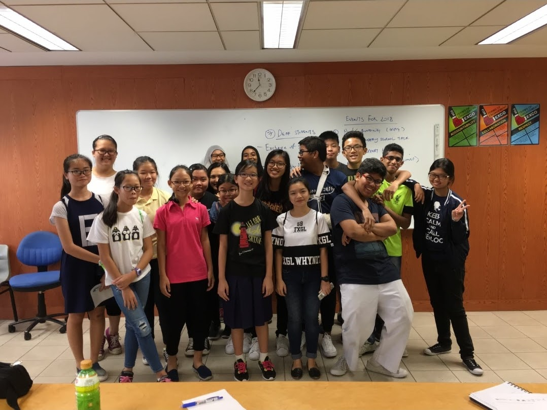 A group of about 20 students posing together in a classroom in front of a whiteboard with a clock above.